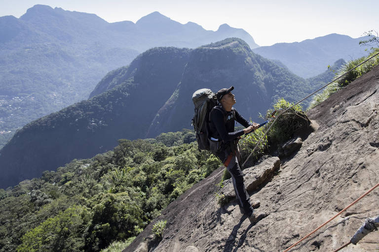 Um escalador está subindo uma rocha íngreme em uma montanha. Ele usa um equipamento de escalada e carrega uma mochila nas costas. Ao fundo, há uma paisagem montanhosa com vegetação densa e várias camadas de montanhas ao longe, sob um céu claro.