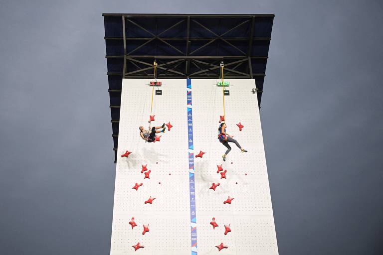 A imagem mostra uma parede de escalada vertical com dois escaladores em ação. Um escalador está à esquerda, enquanto o outro está à direita, ambos utilizando equipamentos de segurança. A parede é coberta com várias agarras vermelhas e possui uma estrutura metálica acima. O céu ao fundo está nublado.
