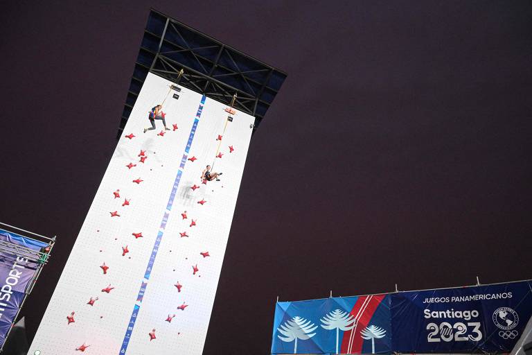 A imagem mostra dois atletas escalando uma parede de escalada vertical, com várias agarras vermelhas. O fundo é escuro, indicando que a competição ocorre à noite. À direita, há um banner com as inscrições 'JUEGOS PANAMERICANOS' e 'Santiago 2023'.