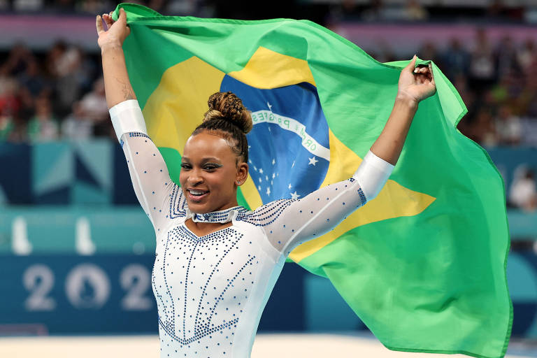 Uma ginasta está em uma competição, levantando a bandeira do Brasil acima da cabeça. Ela usa um traje de competição brilhante, com detalhes em azul e prata. O fundo mostra uma multidão assistindo ao evento.