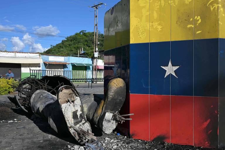 A imagem mostra um monumento danificado que exibe a bandeira da Venezuela. A parte superior do monumento é pintada nas cores da bandeira, com um fundo amarelo, azul e vermelho, e uma estrela branca. Na parte inferior, há uma estrutura de metal retorcida e danificada, que parece ter caído. Ao fundo, é possível ver uma colina e algumas construções coloridas.