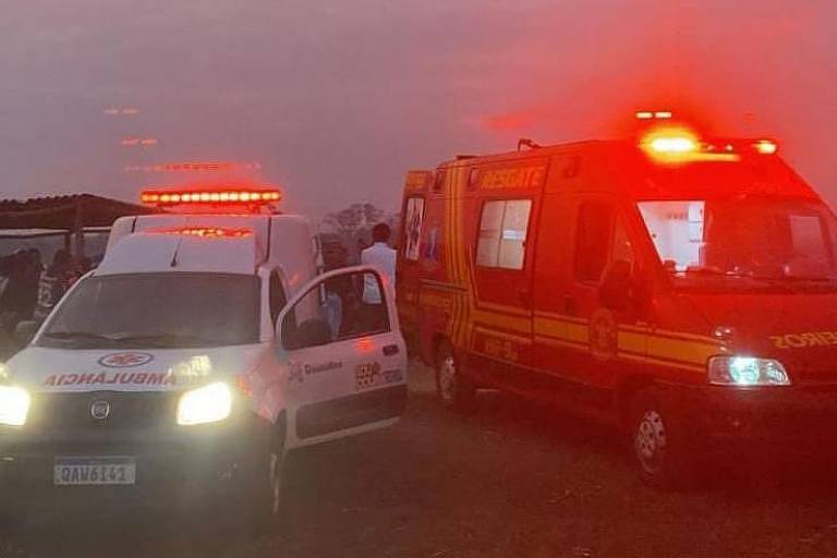 A imagem mostra duas ambulâncias em um local ao ar livre durante o crepúsculo. A ambulância à esquerda é branca com detalhes em vermelho e possui a porta do motorista aberta. A ambulância à direita é vermelha com faixas amarelas e luzes de emergência acesas. Ao fundo, há algumas pessoas e uma estrutura simples coberta. O céu apresenta uma coloração avermelhada devido ao pôr do sol.