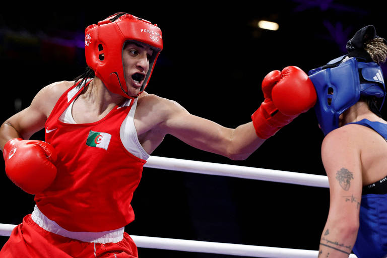 A boxeadora Imane Khelif está com uma roupa vermelha, luvas vermelhas e um protetor facial também vermelho. Mulher branca, está aplicando um golpe com o braço direito contra a rival, também mulher branca. 
