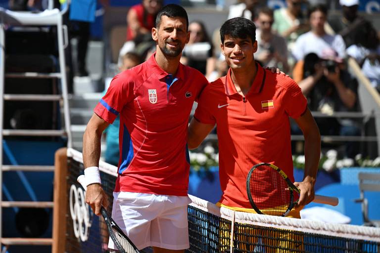 Dois jogadores de tênis posam juntos em um torneio. O jogador à esquerda veste uma camisa vermelha com detalhes azuis e o emblema da Sérvia, enquanto o jogador à direita usa uma camisa vermelha com uma bandeira da Espanha. Ambos seguram raquetes e estão sorrindo, com um fundo de arquibancadas e espectadores.
