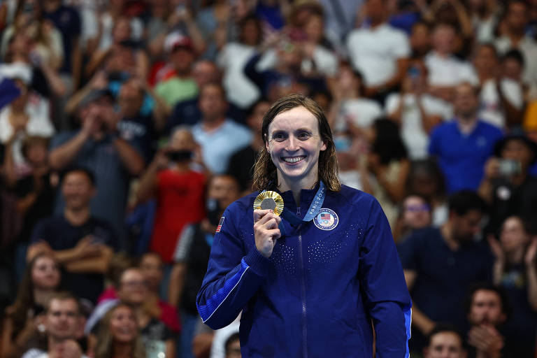 Foto mostra Katie Ledecky da cintura para cima na frente da torcida. A nadadora branca e loira sorri e segura a medalha de ouro com a mão direita. Ela veste um agasalho azul e está com os cabelos molhados.