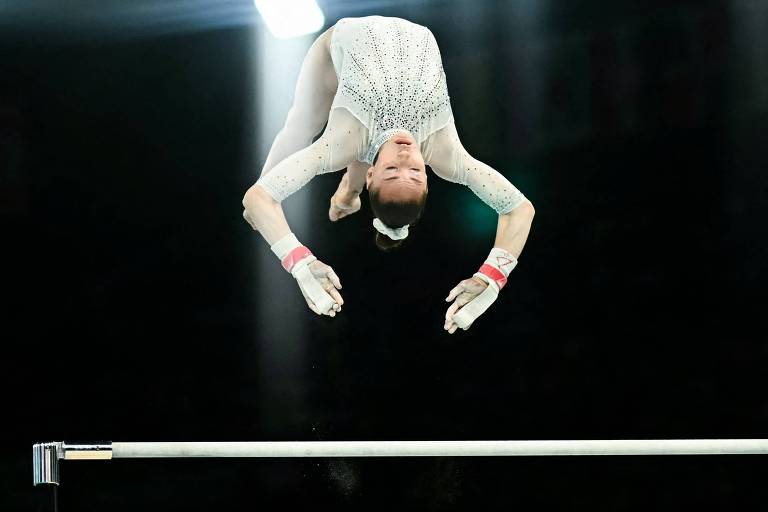 A imagem mostra uma ginasta realizando um salto sobre uma barra, com o corpo em posição invertida. O fundo é escuro, com luzes brilhantes que criam um efeito dramático. A ginasta está vestido com um traje de competição e usa luvas brancas.