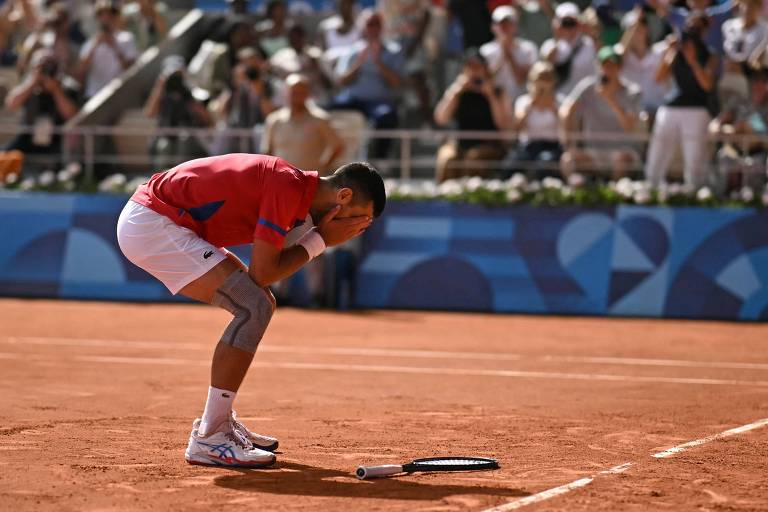 Um jogador de tênis, vestido com uma camiseta vermelha e shorts brancos, está se curvando sobre sua raquete no chão, demonstrando uma forte emoção. Ao fundo, uma multidão de espectadores observa, alguns aplaudindo. O cenário é um campo de tênis de terra batida, sob um céu claro.