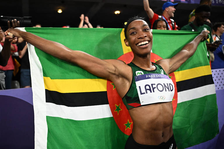 Foto mostra aa atleta negra retinta com a bandeira de seu país erquida pelas suas mãos atrás das suas costas. Lafond está sorrindo e veste um top verde com seu nome escrito nele. 