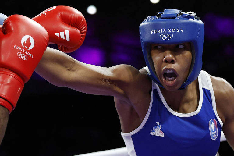 A imagem mostra uma atleta de boxe em ação, usando um uniforme azul e um capacete azul com a inscrição 'PARIS 2024'. Ela está desferindo um golpe com a mão direita, que está coberta por uma luva vermelha com o logotipo dos Jogos Olímpicos de Paris 2024. O fundo é desfocado, com luzes que sugerem um ambiente de competição.
