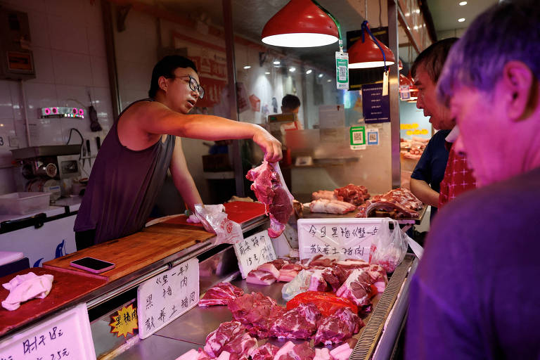 Fotografia mostra uma banca de rua com carne de porco exposta. Há um homem de regata cinza segurando um pedaço de carne, atrás da bancada. Outros dois homens, desfocados, observam o balcão.