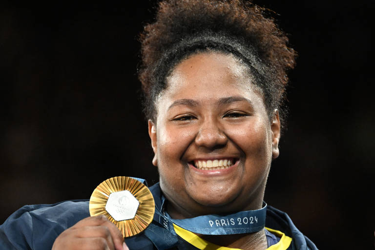 Beatriz, mulher preta, está sorrindo. Tem os cabelos presos no alto e segura com a mão direita sua medalha de ouro. Fundo preto.