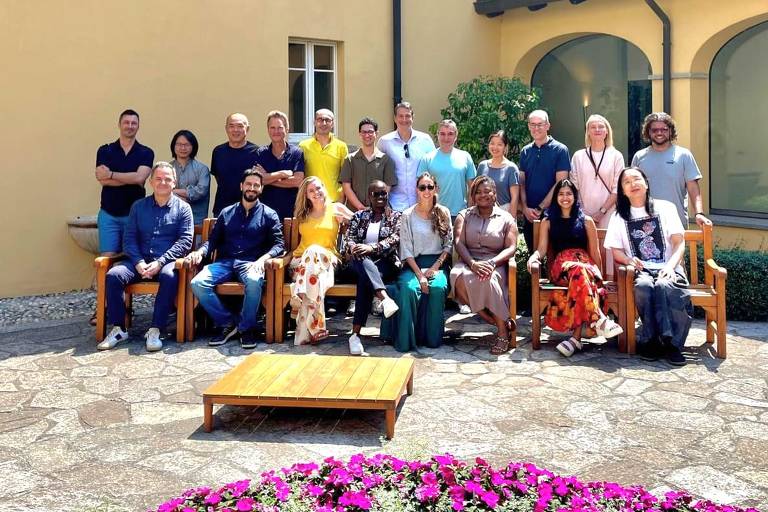 A imagem mostra um grupo de aproximadamente 20 pessoas reunidas em um ambiente externo, posando para a foto. Elas estão sentadas e em pé em um espaço com piso de pedras, cercadas por um jardim com flores roxas. Ao fundo, há uma parede amarela e janelas. Algumas pessoas estão segurando objetos, e um cachorro está presente na frente do grupo.