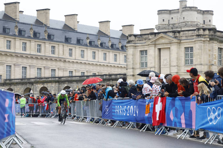 A imagem mostra um ciclista em uma competição de ciclismo durante os Jogos Olímpicos de Paris 2024. O ciclista está vestido com um traje verde e pedala em uma estrada molhada, enquanto uma multidão de espectadores assiste ao evento. Algumas pessoas seguram guarda-chuvas, e há bandeiras visíveis, incluindo uma da Suíça. Ao fundo, um edifício histórico é visível, com uma arquitetura clássica.
