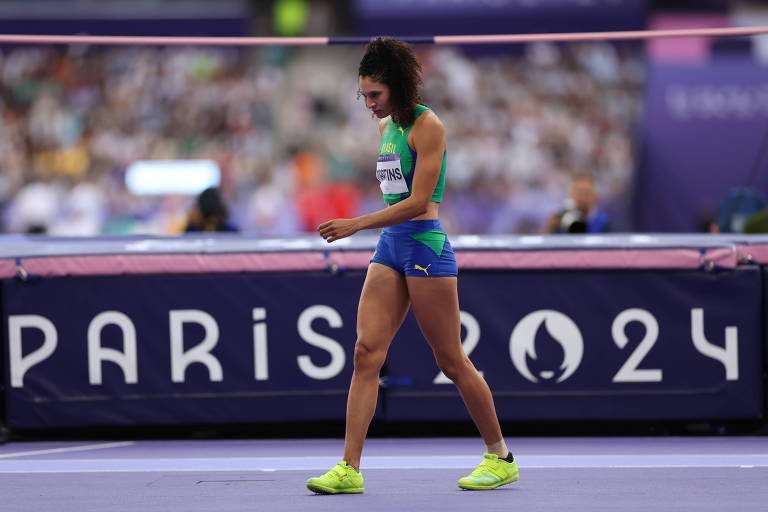 Uma atleta está caminhando na pista de atletismo, vestindo um uniforme azul e verde, com tênis amarelos. Ao fundo, há uma multidão assistindo ao evento. O painel exibe 'PARIS 2024'.
