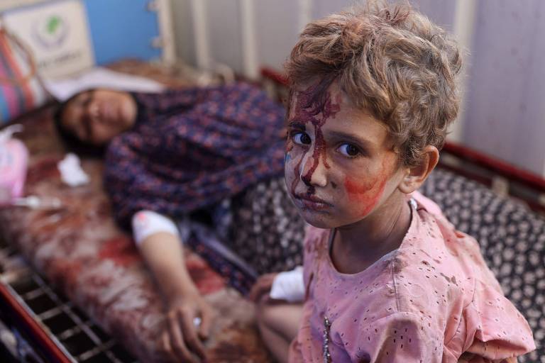 A imagem mostra uma criança com cabelo curto e bagunçado, olhando diretamente para a câmera. Seu rosto está sujo, com marcas de sangue. Ao fundo, uma mulher inconsciente deitada em uma cama.