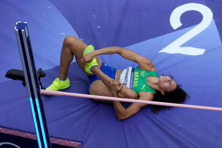 A imagem mostra uma atleta de salto com vara caída sobre o colchão de aterrissagem, segurando a perna. Ela está vestindo um uniforme verde e amarelo, com tênis amarelos. O varal está visivelmente acima dela, e o número '2' está marcado no fundo.
