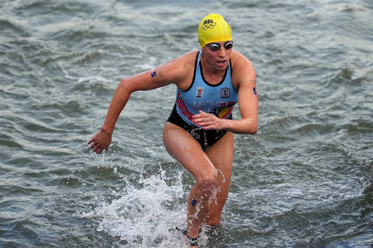 Uma atleta está saindo da água durante uma competição de natação. Ela usa um maiô de competição azul e amarelo, com uma touca amarela. A água está salpicando ao seu redor enquanto ela corre em direção à margem.