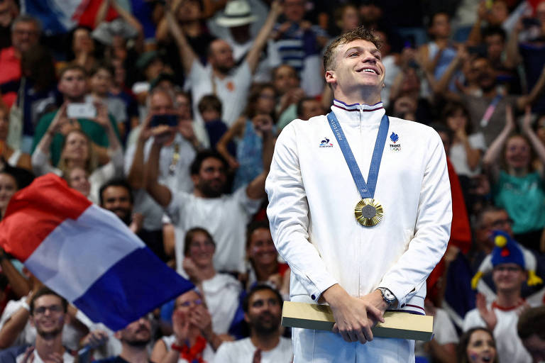 Com a medalha de ouro da prova dos 200 m peito nos Jogos Olímpicos de Paris pendurada em torno do pescoço, o nadador francês Léon Marchand sorri; atrás dele está a torcida francesa