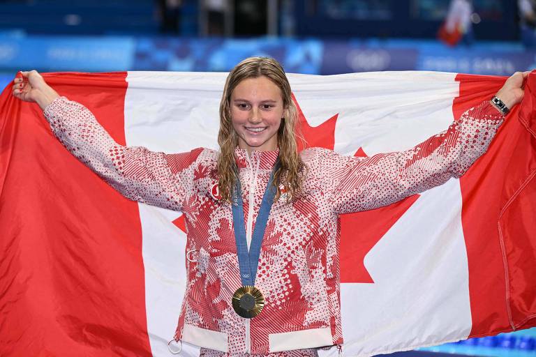 A nadadora Summer Mcintosh festeja o ouro nos 200 m medley nas Olimpíadas de Paris sorrindo, com a medalha no peito e segurando a bandeira do Canadá