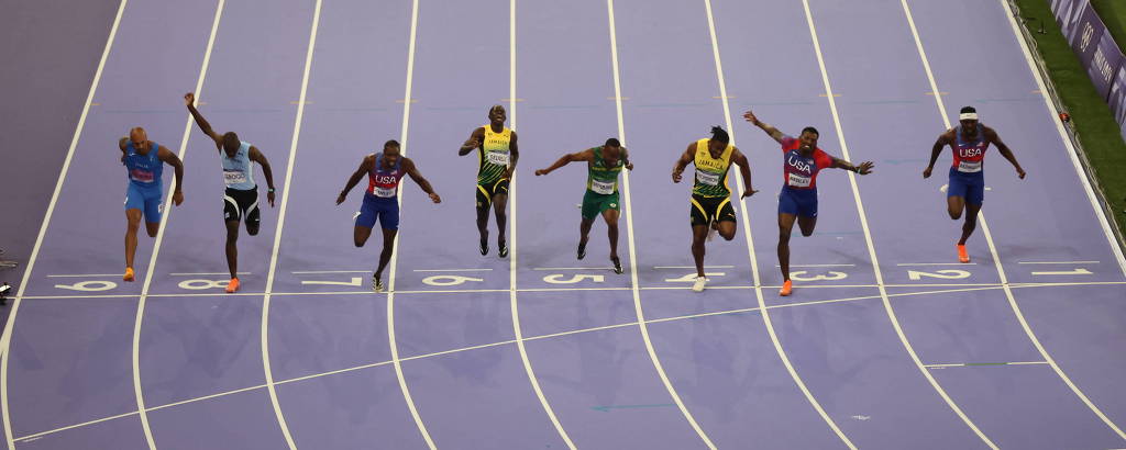A imagem mostra uma corrida de atletismo com sete atletas competindo em uma pista de corrida. Os corredores estão em diferentes posições, alguns com os braços levantados, enquanto outros estão em movimento. A pista é de cor azul e as linhas brancas delimitam as raias. O ambiente parece ser um estádio, com grama ao fundo.