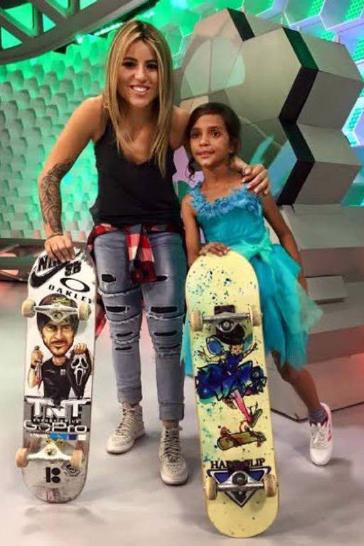 Uma mulher loira posa com criança vestida de fada e skate nas mãos