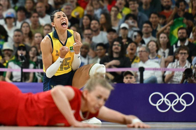 Uma jogadora de vôlei, vestindo um uniforme amarelo e azul, está em uma posição de celebração, com os braços levantados e uma expressão de alegria. Ao fundo, uma multidão de torcedores pode ser vista, enquanto uma jogadora adversária está caída no chão, em um uniforme vermelho.
