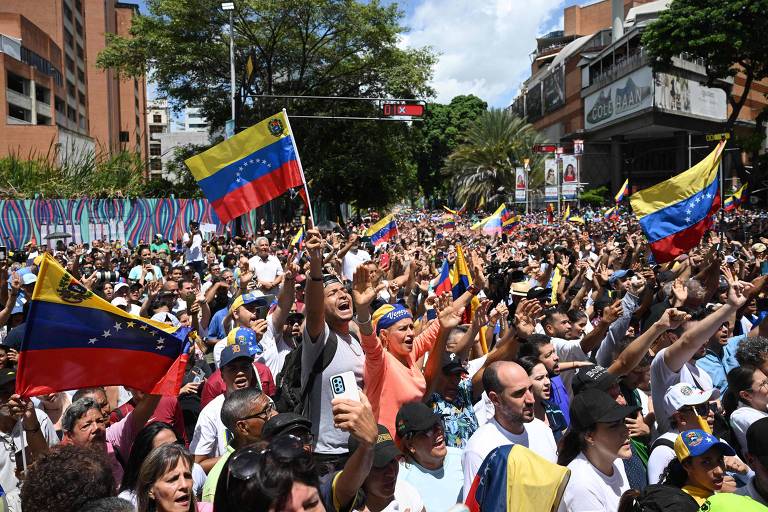 A imagem mostra uma grande multidão reunida em uma manifestação nas ruas, com pessoas segurando bandeiras da Venezuela. O céu está claro e há prédios ao fundo. A atmosfera parece ser de entusiasmo e protesto.
