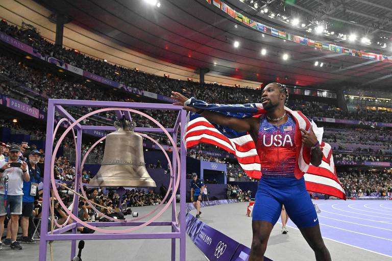 Um atleta está tocando um sino em um estádio, vestindo uma camisa com a inscrição 'USA' e segurando uma bandeira dos Estados Unidos. O ambiente é de uma competição esportiva, com uma grande multidão ao fundo
