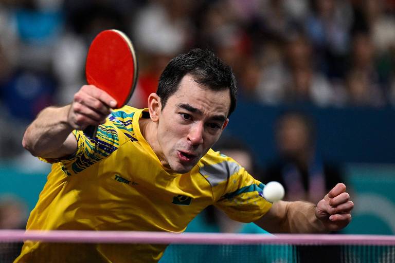 O mesa-tenista brasileiro Hugo Calderano, usando raquete com borracha vermelha, rebate a bolinha no jogo em que perdeu o bronze olímpico em Paris para o francês Félix Lebrun