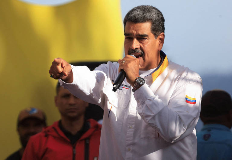 Um homem de pele clara, com cabelo escuro e bigode, está falando em um microfone. Ele usa uma camisa branca com detalhes em amarelo e a bandeira da Venezuela. Ao fundo, há pessoas e uma grande estrutura amarela. O homem gesticulacom a mão direita.