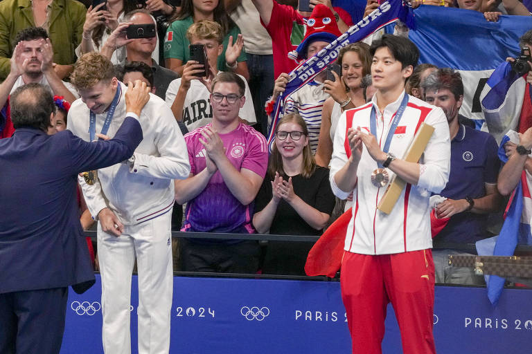 A imagem mostra uma cerimônia de premiação olímpica, onde um atleta está recebendo uma medalha de ouro de um oficial, enquanto outro atleta, ao lado, segura uma medalha de bronze. O público ao fundo está aplaudindo e alguns estão segurando celulares para registrar o momento. O ambiente é festivo, com bandeiras e pessoas vestindo camisetas de diferentes países.