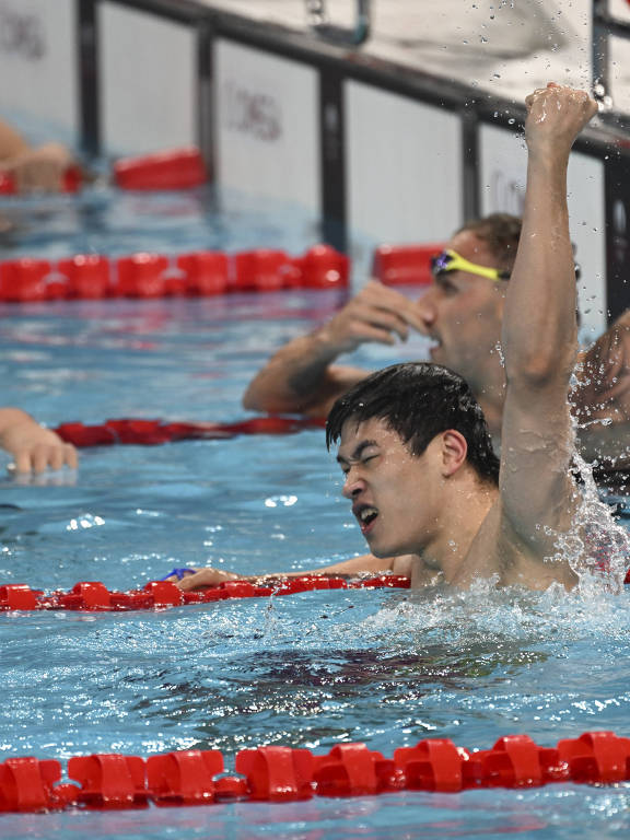 A imagem mostra um atleta de natação comemorando após uma prova. Ele está na piscina, com a mão levantada em sinal de vitória, enquanto água espirra ao seu redor. Ao fundo, outros nadadores também estão na borda da piscina, alguns com óculos de natação. O ambiente é de competição, com as marcas de raia visíveis.