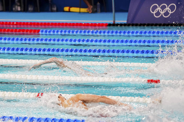 A imagem mostra uma competição de natação em uma piscina olímpica. Vários nadadores estão em ação, com um nadador em primeiro plano, parcialmente submerso, e outro nadador ao fundo. As raias da piscina são demarcadas por cordas brancas e azuis, e há respingos de água visíveis. No fundo, pode-se ver uma estrutura com o logotipo dos Jogos Olímpicos.