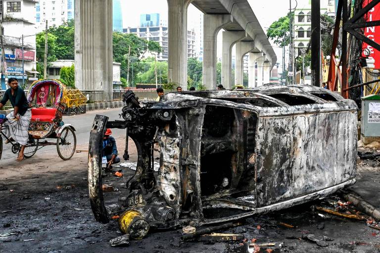 Fotografia mostra utilitário (aparentemente uma van) incendiada. Ao fundo, uma mulher pedala. 