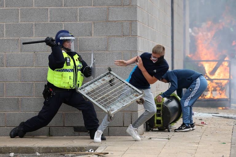 A imagem mostra um policial em uniforme, segurando um bastão, enquanto tenta conter um jovem que está se afastando, segurando uma grade. Ao fundo, há uma grande chama, indicando um incêndio. Outro jovem está próximo, observando a cena. O ambiente parece ser urbano, com paredes de bloco de concreto e detritos no chão.