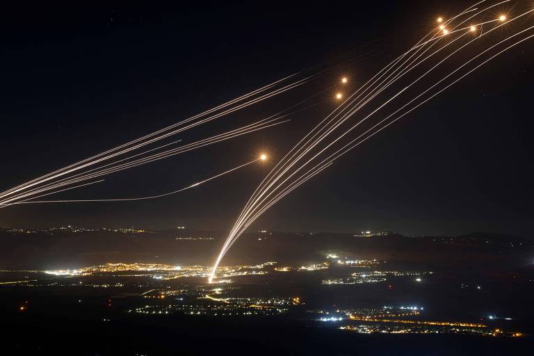 Numa noite escura, riscos no céu indicam trajetória de foguetes de defesa que explodem contra foguetes que foram lançados contra Israel, em bolas de fogo. Há uma cidade iluminada na paisagem