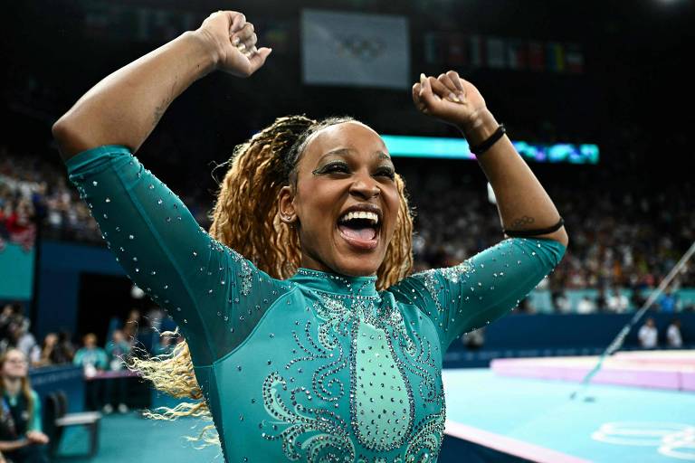 Uma atleta está comemorando com os braços levantados, sorrindo amplamente. Ela usa um traje de ginástica verde com detalhes brilhantes. Ao fundo, há uma multidão e bandeiras, indicando que o evento é de grande importância, possivelmente uma competição olímpica.
