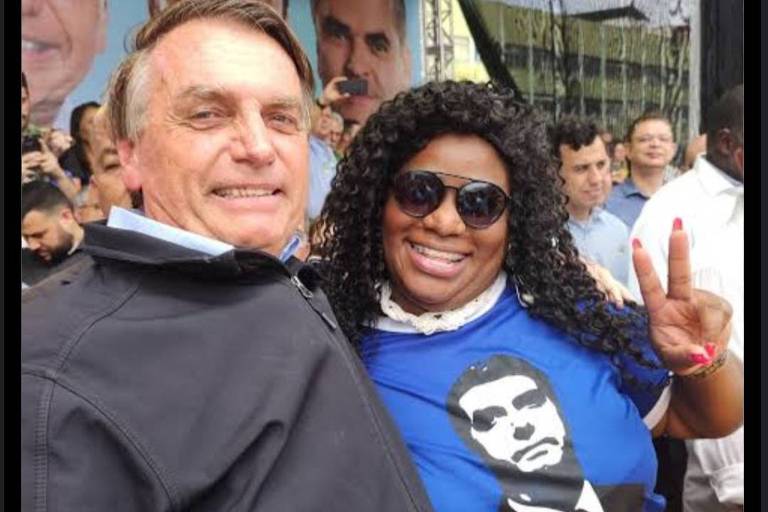 Duas pessoas posam para foto. A da esquerda é Jair Bolsonaro, homem branco, de jaqueta escura. A seu lado está uma mulher preta, de óculos escuros e camiseta azul com ilustração de Bolsonaro e escrito "Bolsonaro presidente". 