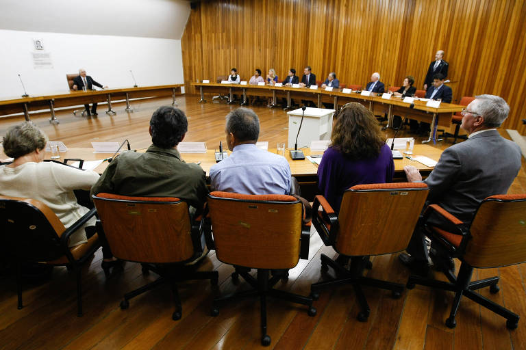 mesa oval, com integrantes sentados em torno dela. Paredes são de madeira