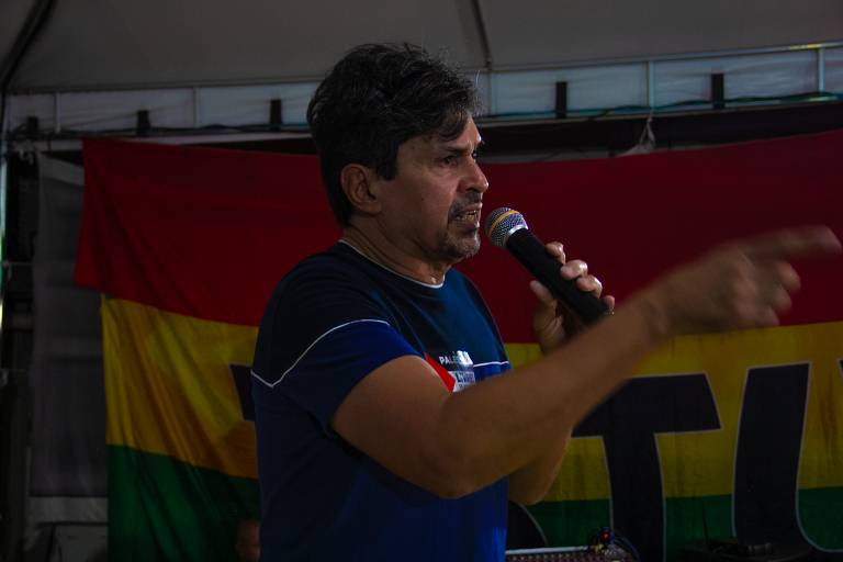 Um homem de cabelo escuro e curto, com uma camiseta azul, está falando em um microfone. Ele gesticula com a mão direita enquanto se expressa. Ao fundo, há uma bandeira com as cores vermelho, amarelo e verde.