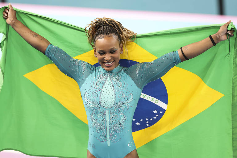 Sorrindo e usando collant azul, a ginasta Rebeca Andrade comemora com a bandeira do Brasil o ouro no solo nas Olimpíadas de Paris