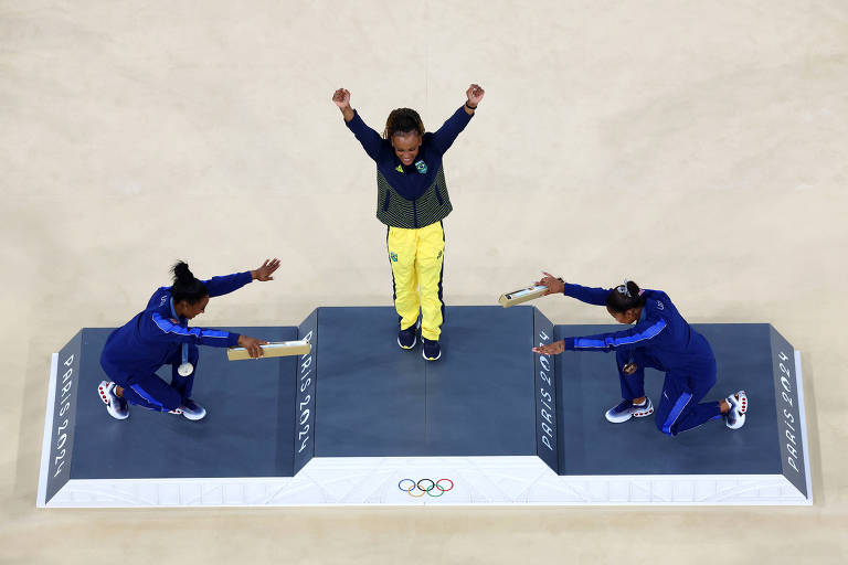 A imagem mostra uma cerimônia de premiação olímpica. No centro, um atleta está em pé no pódio, levantando os braços em sinal de vitória. Ele usa uma roupa amarela e cinza. À esquerda e à direita, dois outros atletas estão agachados, segurando bastões. Todos estão em um pódio com o logotipo dos Jogos Olímpicos visível.
