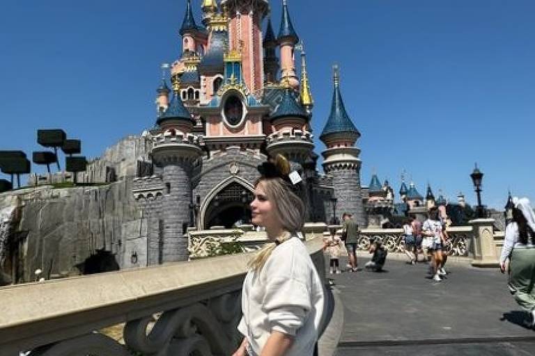 Em foto colorida, mulher de blusa e saia branca posa para foto em frente a uma castelo