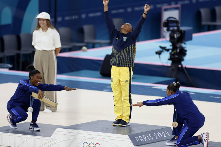 A imagem mostra uma cerimônia de premiação olímpica. No centro, uma atleta vestindo um uniforme amarelo e azul levanta os braços em celebração. À esquerda, uma atleta com uma medalha de prata sorri e se agacha, enquanto à direita, outra atleta se ajoelha, estendendo a mão. Ao fundo, uma mulher com um chapéu branco observa. O cenário é decorado com elementos olímpicos e a inscrição 'PARIS 2024'.
