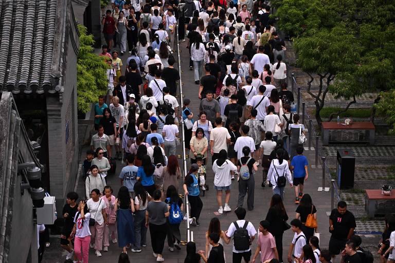 A imagem mostra uma grande multidão de pessoas caminhando em um caminho estreito. As pessoas estão vestidas em roupas variadas, predominantemente em tons claros e escuros. Há árvores ao longo do caminho e edifícios visíveis nas laterais. A perspectiva é de cima, proporcionando uma visão ampla da movimentação.