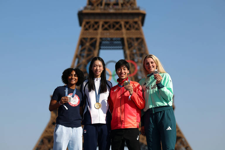 Quatro pessoas posam para a foto em um palco ao ar livre, com a Torre Eiffel visível ao fundo. O céu está claro e azul. As pessoas estão vestidas de forma casual, algumas fazendo gestos de vitória com as mãos.
