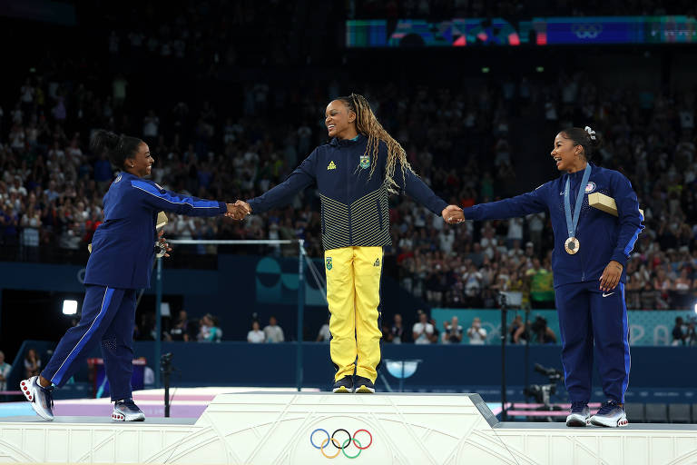 Três atletas estão em um pódio olímpico. No centro, uma atleta vestindo um uniforme amarelo e verde está recebendo as mãos de duas atletas, uma à esquerda vestindo um uniforme azul e outra à direita com um uniforme azul escuro e uma medalha ao redor do pescoço. O fundo mostra uma multidão assistindo ao evento.