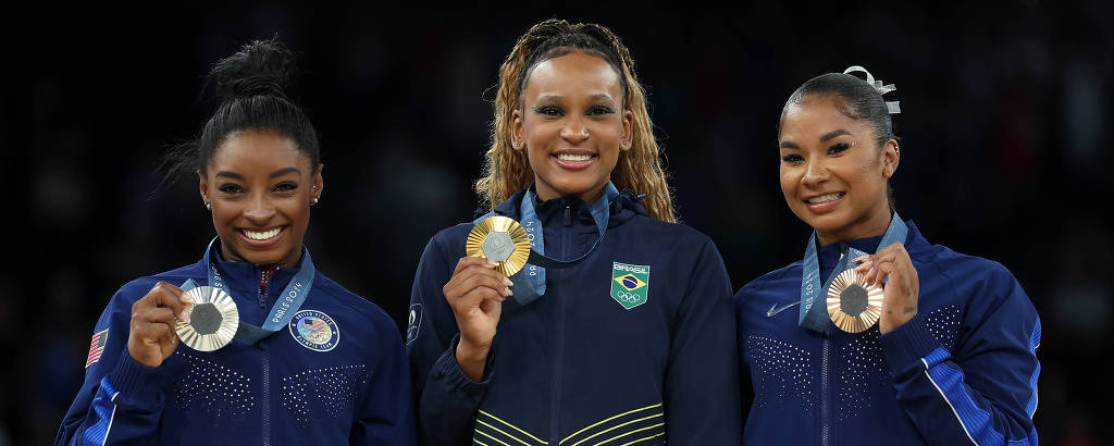 Três atletas estão no pódio, segurando suas medalhas. A atleta do meio, que usa um uniforme amarelo e verde, exibe uma medalha de ouro. As outras duas atletas, à esquerda e à direita, usam uniformes escuros e seguram medalhas de prata e bronze, respectivamente. O fundo é desfocado, sugerindo um ambiente de competição.