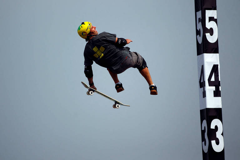 Um skatista está realizando um salto acrobático no ar, segurando seu skate. Ele usa um capacete amarelo e uma camiseta cinza, além de joelheiras e cotoveleiras. Ao fundo, há uma estrutura com números que parecem indicar alturas, com os números 33, 44 e 55 visíveis.
