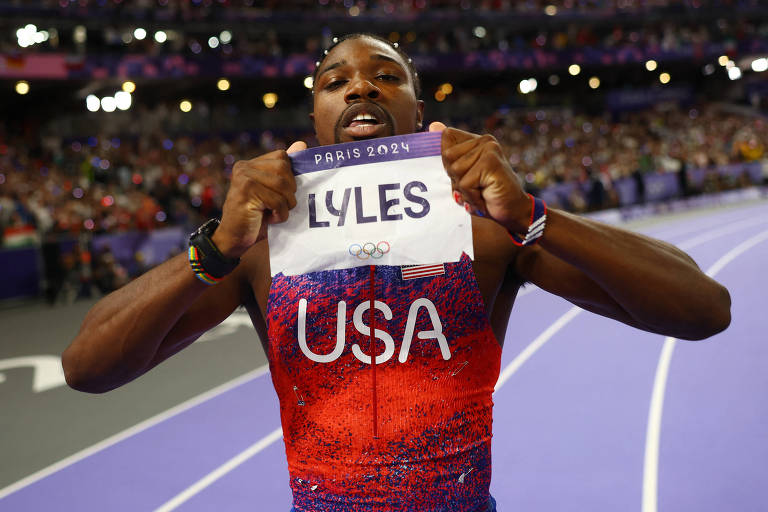 Noah Lyles, velocista dos EUA, está posando em uma pista de atletismo, segurando uma placa com o nome 'LYLES' e a sigla 'USA'. O atleta está vestido com um uniforme vermelho e azul, e há uma multidão ao fundo.